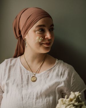 A person in white clothing and brown headscarf poses with natural light casting soft shadows.
