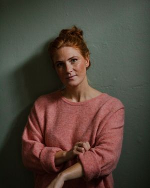 A person in pink sweater poses against a gray wall with natural lighting.