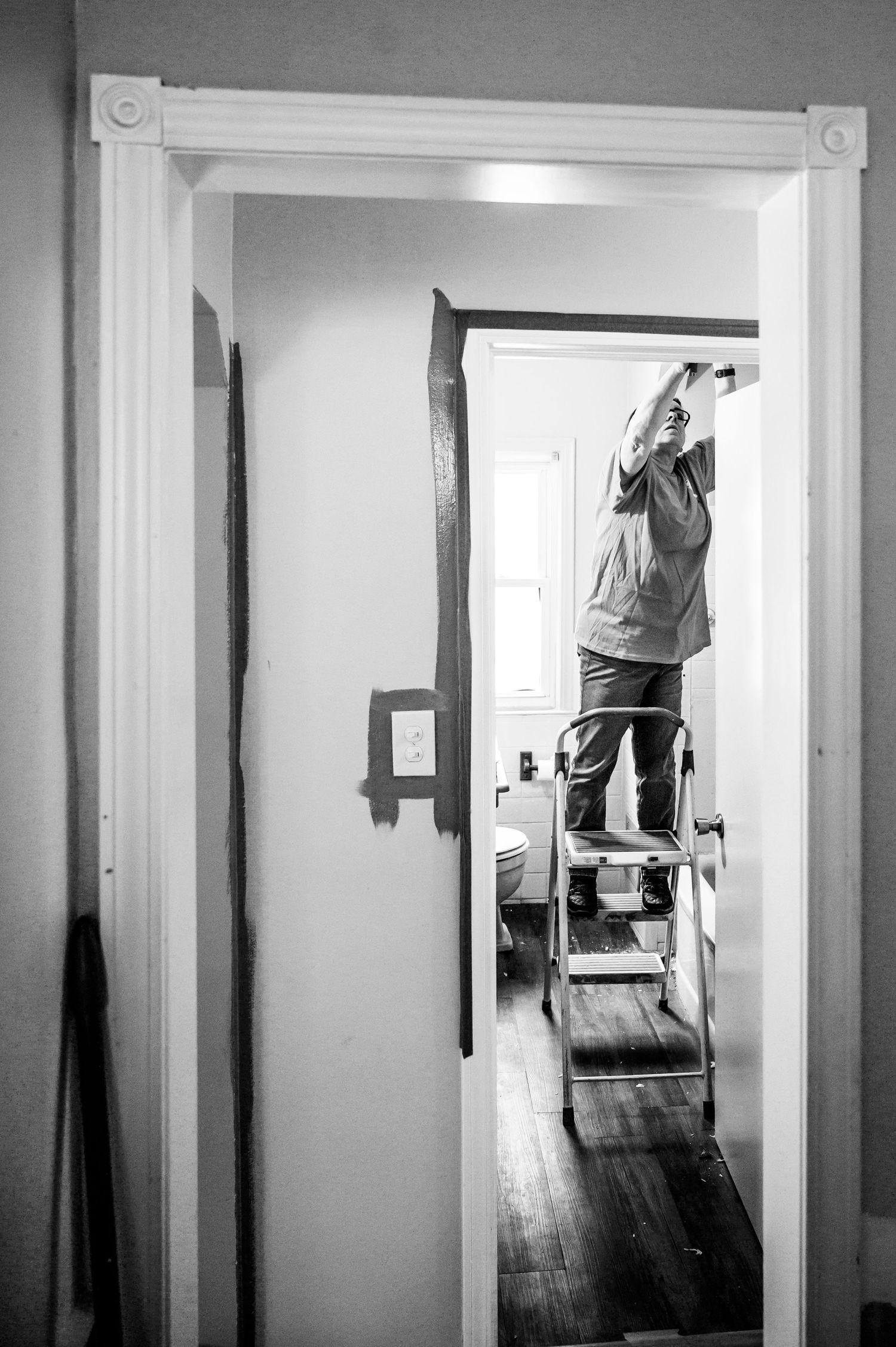 Black and white image of someone standing on ladder painting interior doorway trim.