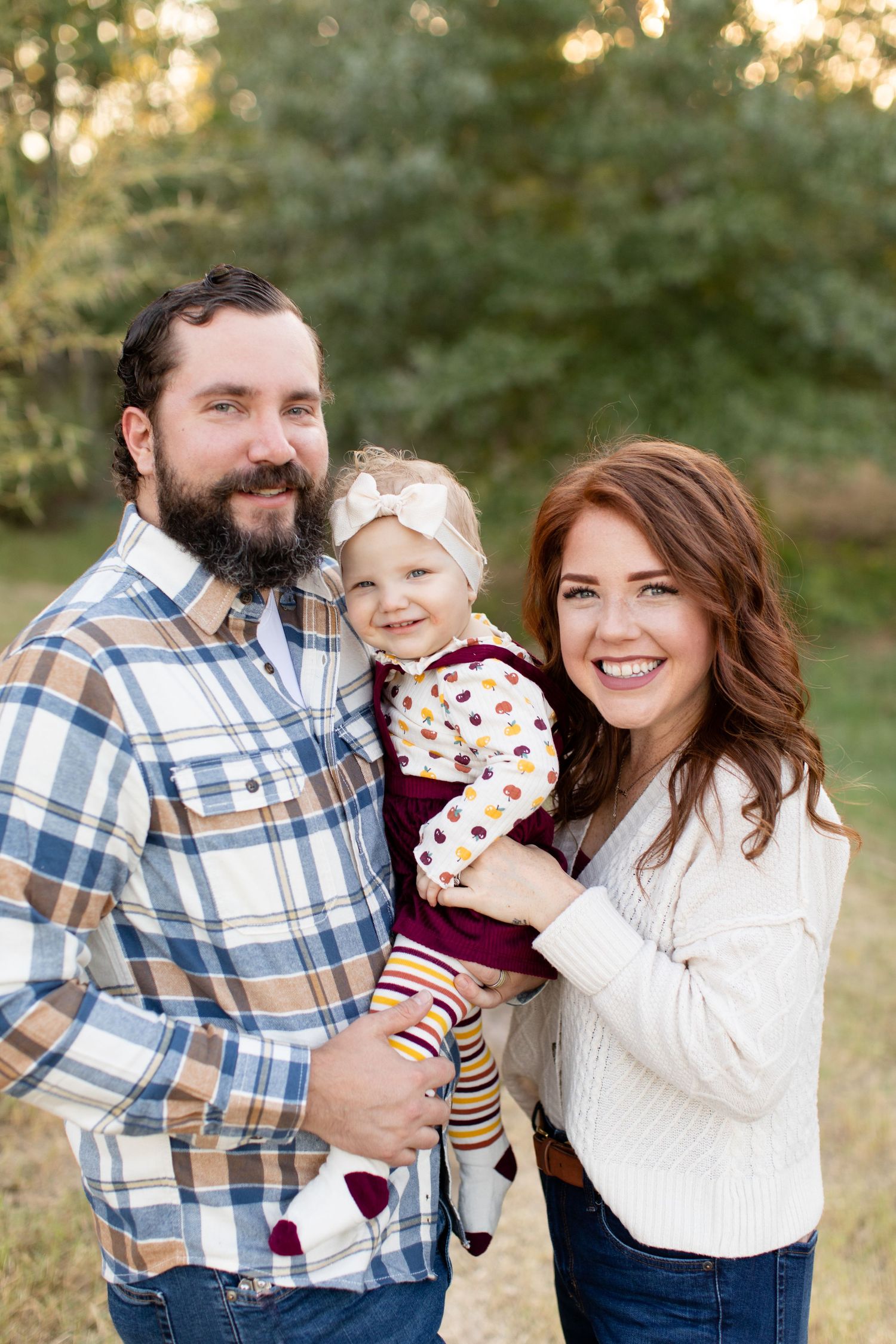 Family outdoor portrait featuring couple in casual fall attire with small child in burgundy dress and striped leggings.