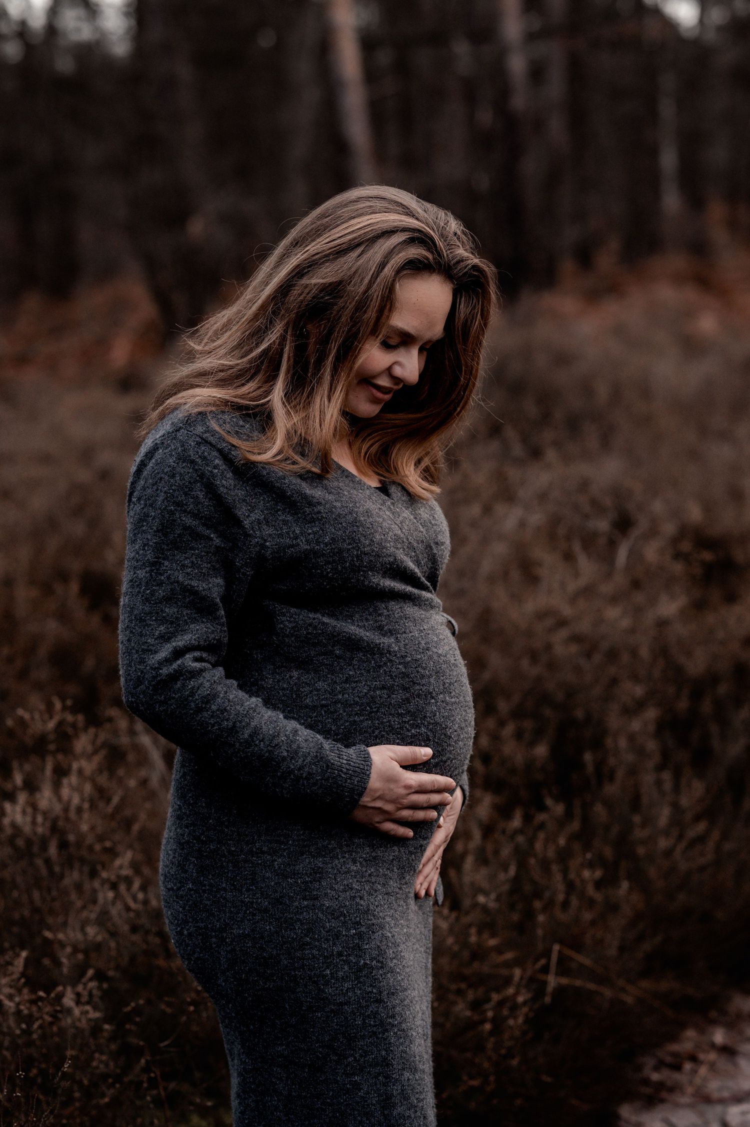 Maternity photos of woman in dark sweater posing against moody forest backdrop.
