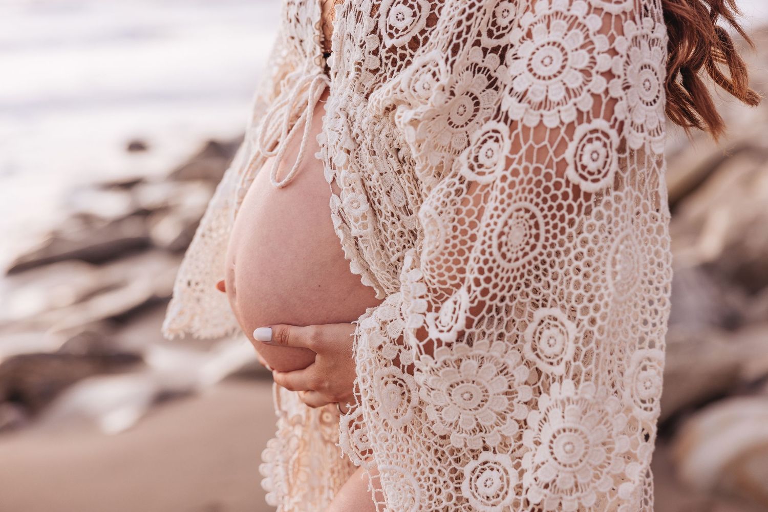 Delicate lace details of a sheer white maternity dress shown in close-up on the beach.