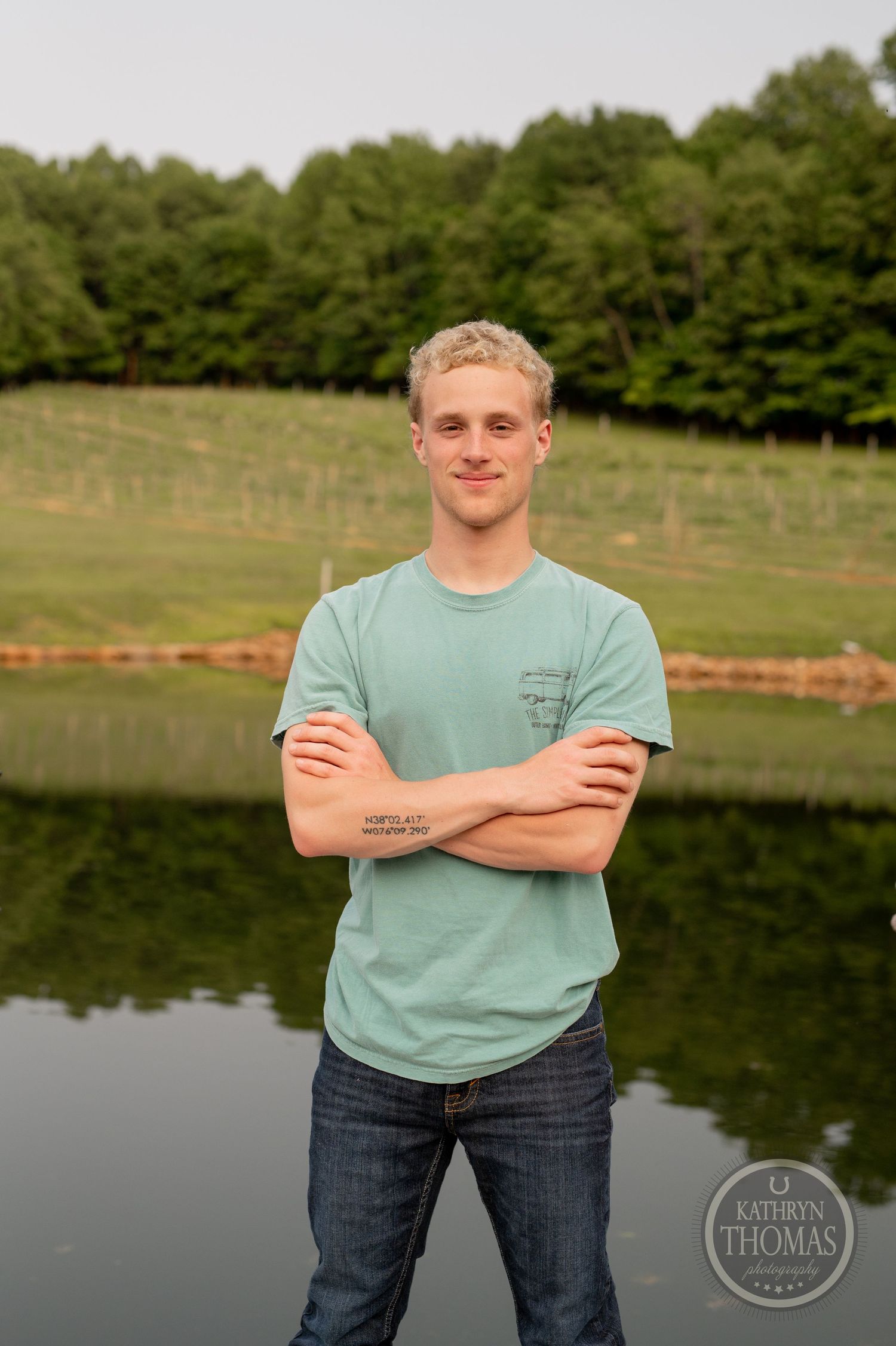 Standing in front of a serene pond with trees, wearing a mint green t-shirt and jeans on a summer evening.