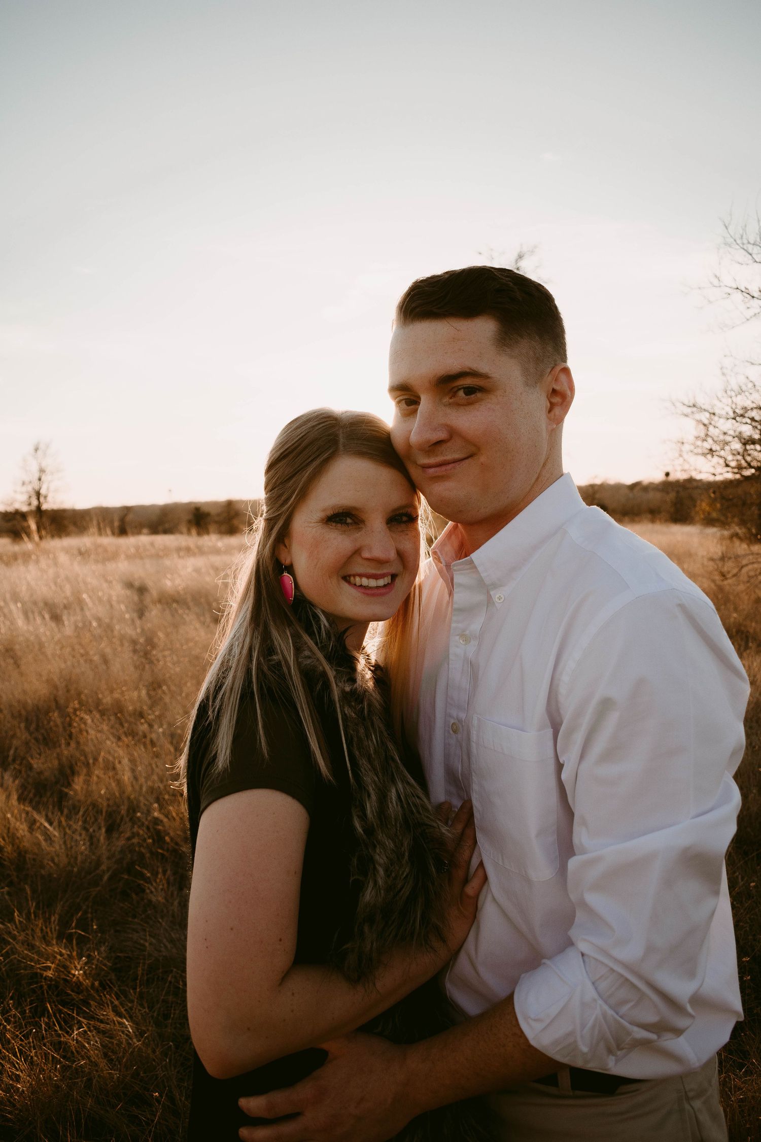 A couple poses together in warm evening light with a natural field backdrop.
