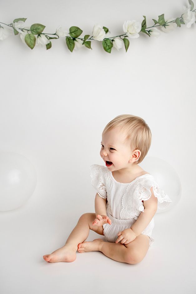 One year old girl in white outfit laughing at white balloons.