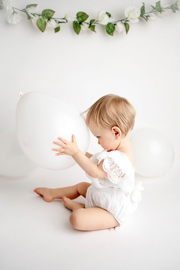 One year old girl holding a white balloon to her forehead.