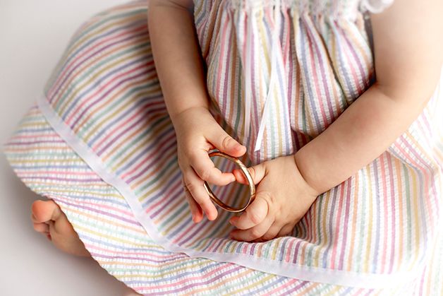 One year old girl in striped dress, sitting on white backdrop with gold bracelet in hands.