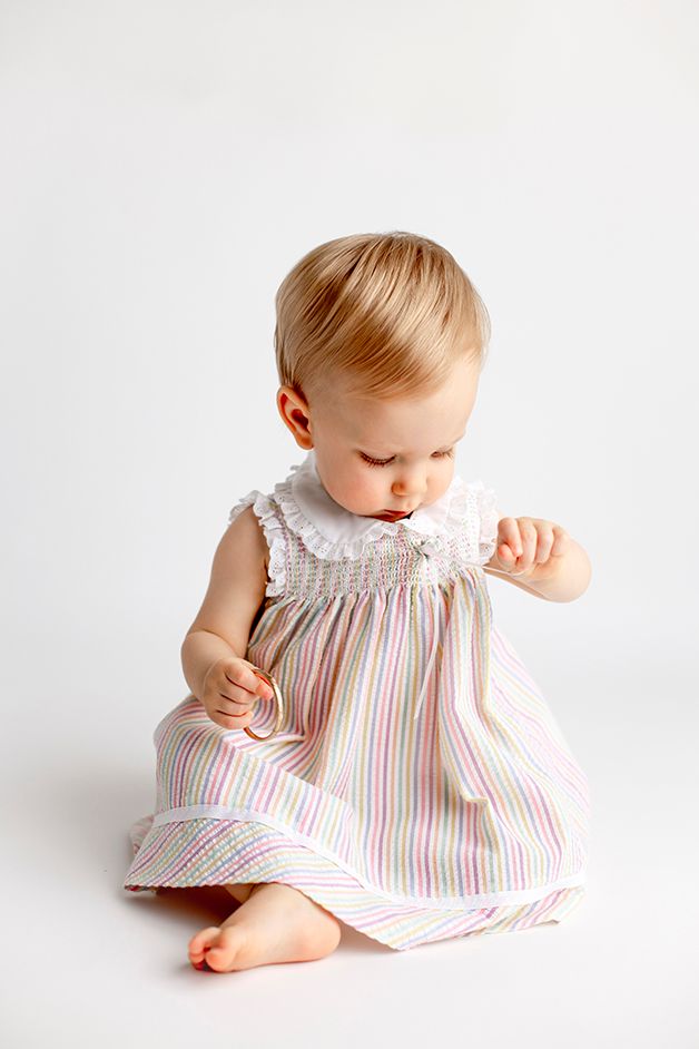 One year old girl in striped dress looking and pulling at ribbons on her dress.