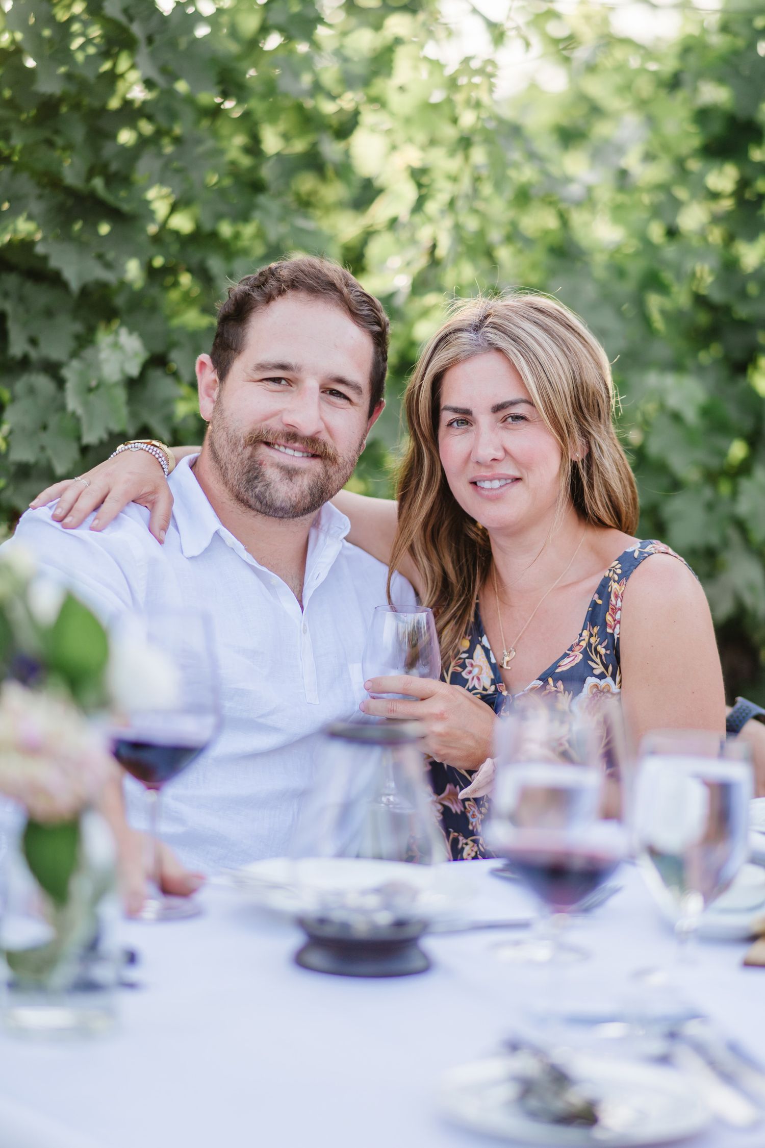 A couple smiles and laughs together at an outdoor dining table with wine glasses.