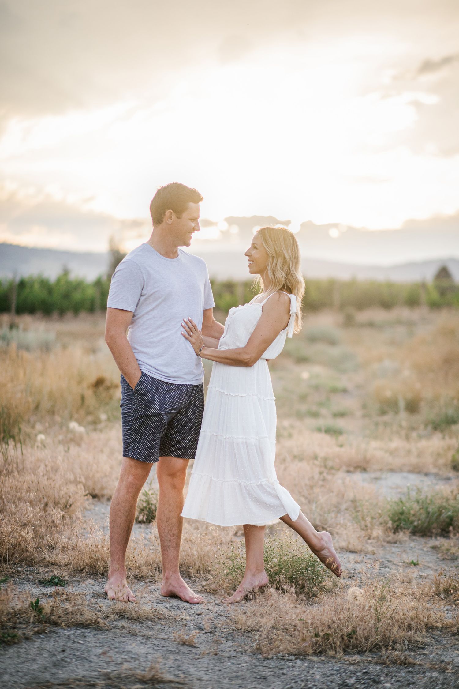 A couple in casual summer outfits dances together in a golden field at sunset.