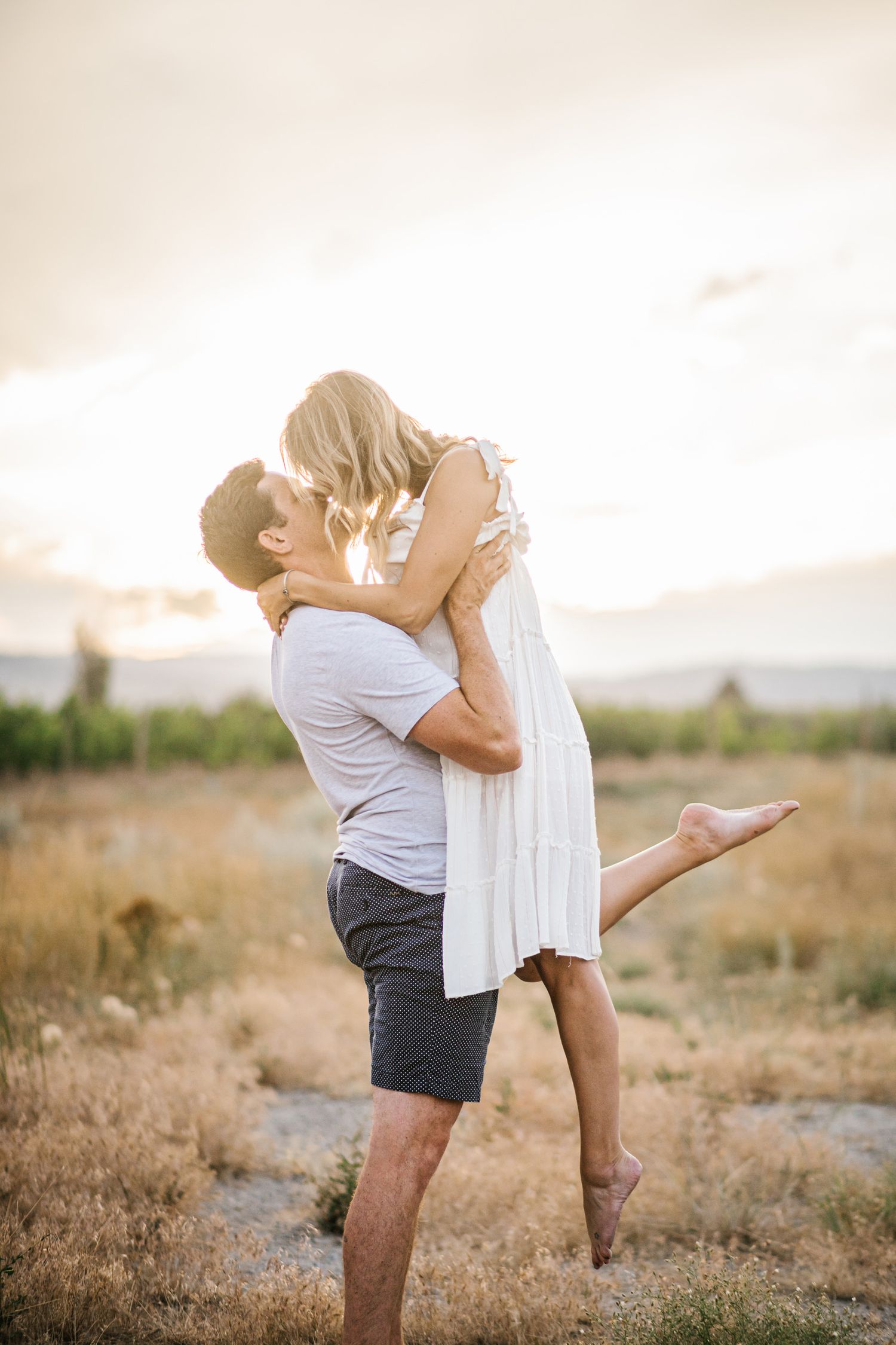 A romantic couple embraces and lifts each other in a desert field during sunset.