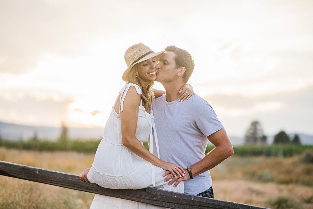 A couple shares romantic moments while sitting on a fence at sunset.