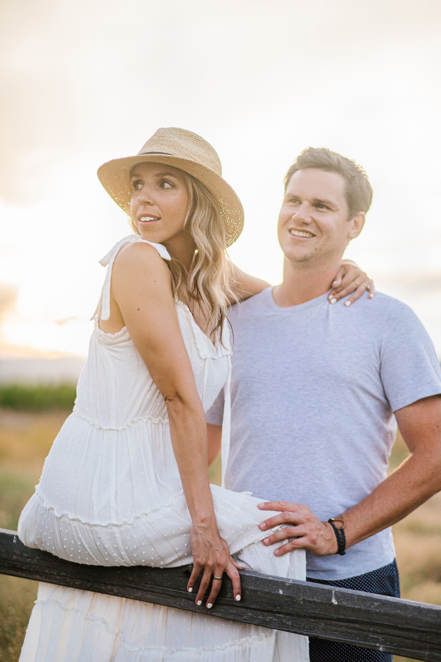 Couple in casual attire sharing a romantic moment while leaning on a wooden fence at sunset.