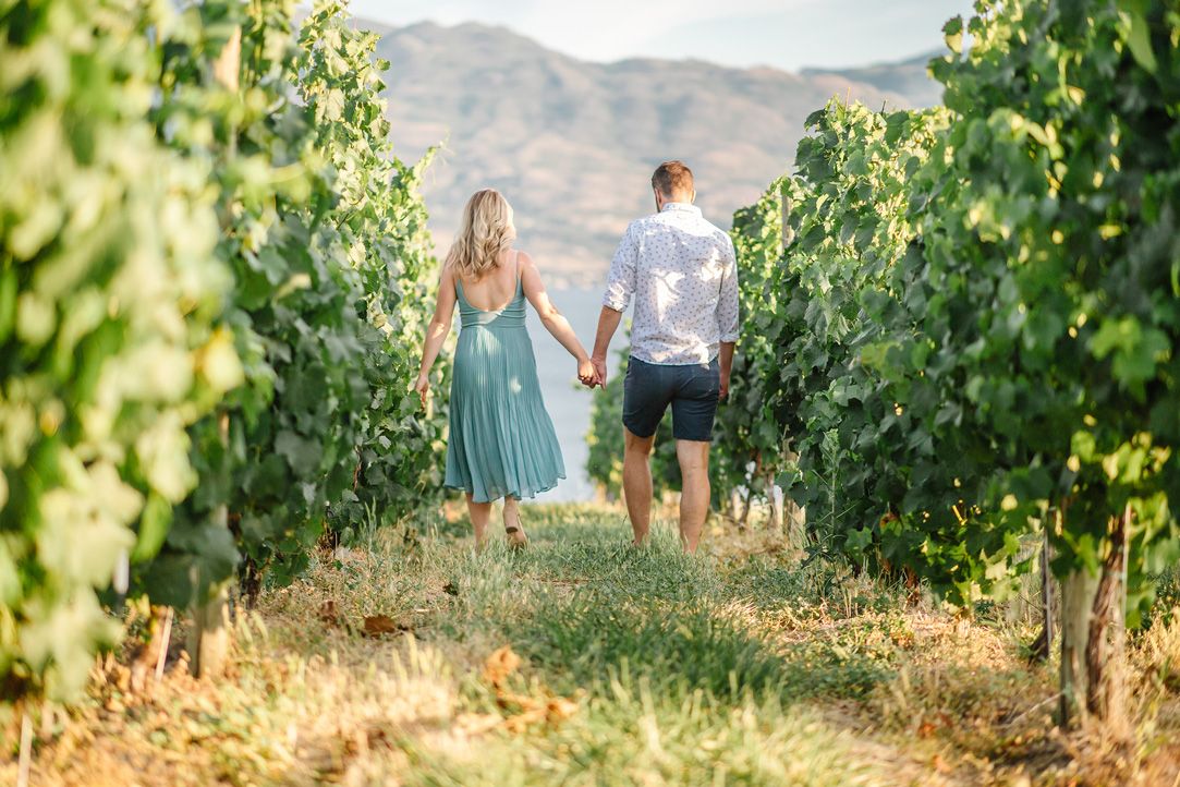 Couple walking through lush vineyard with mountain landscape in background.