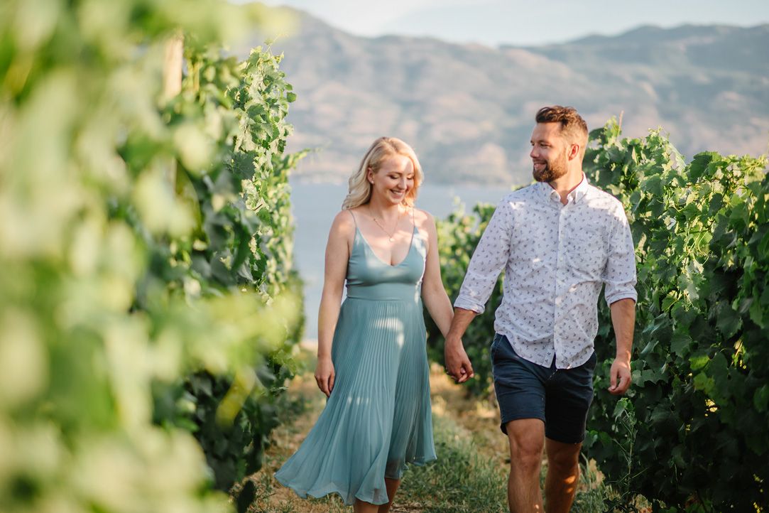 Couple strolling through green vineyard rows with mountains visible in background.