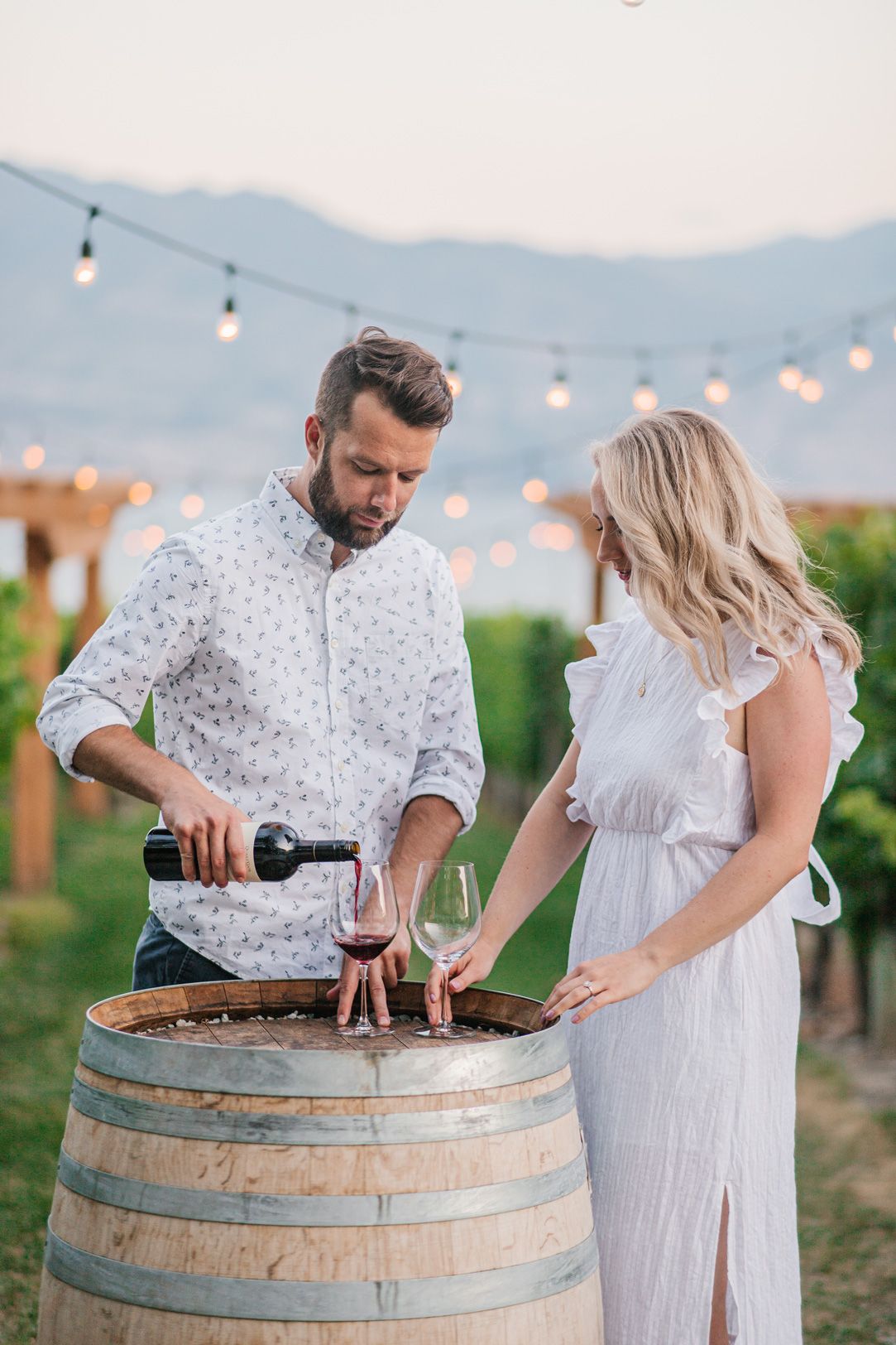 Couple pouring wine at rustic barrel in vineyard with string lights overhead.