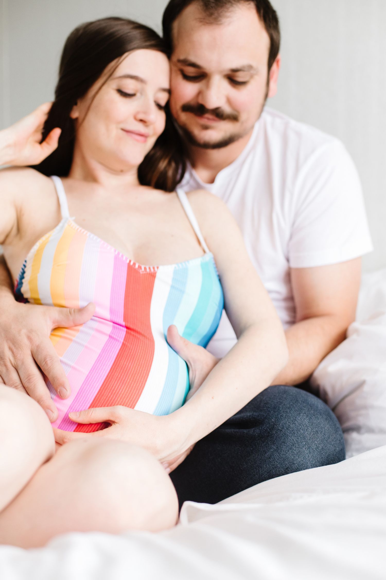 Couple shares intimate moment on white bedding, with person wearing colorful striped summer dress.