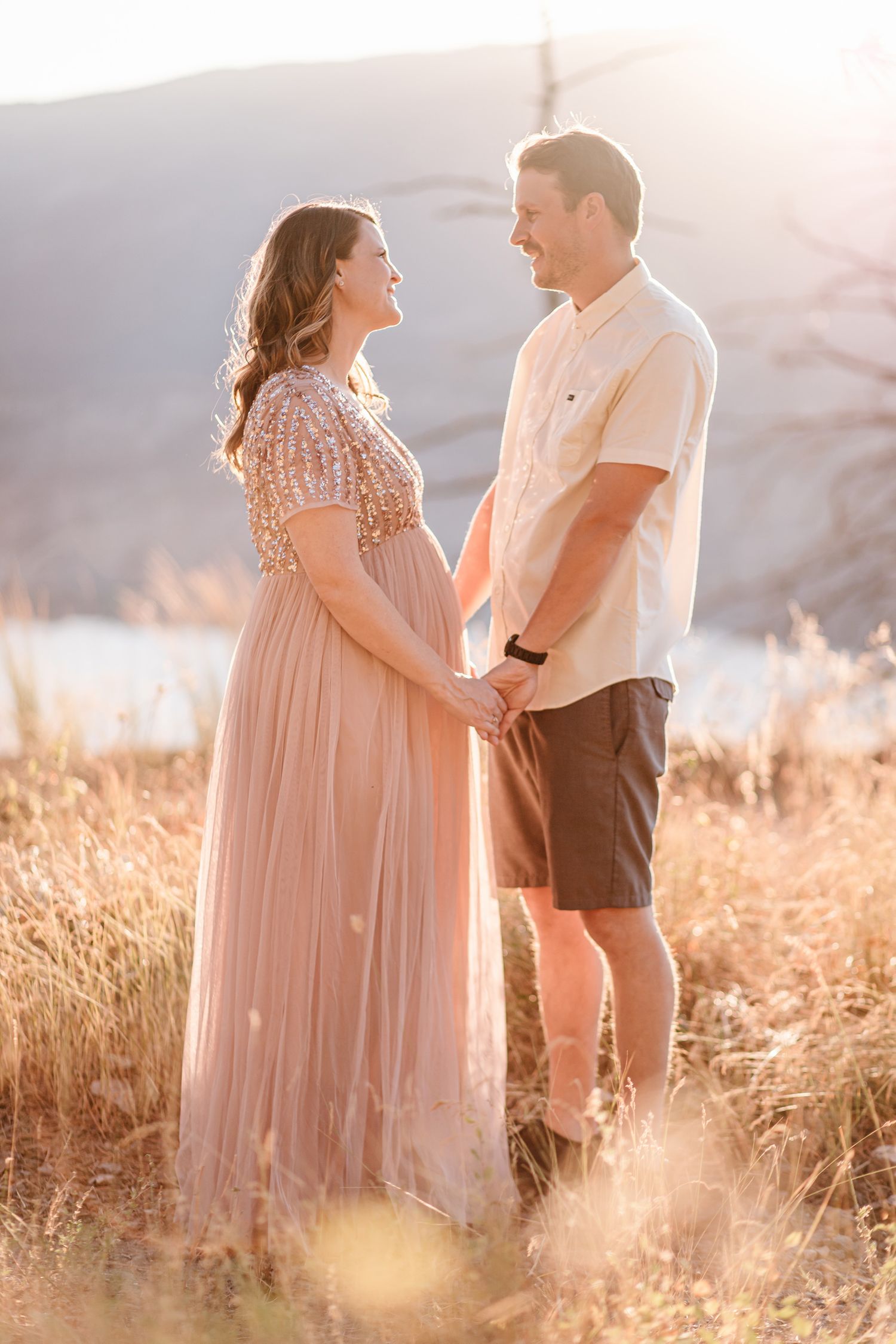 A couple stands together in tall golden grass during a sunset maternity session.