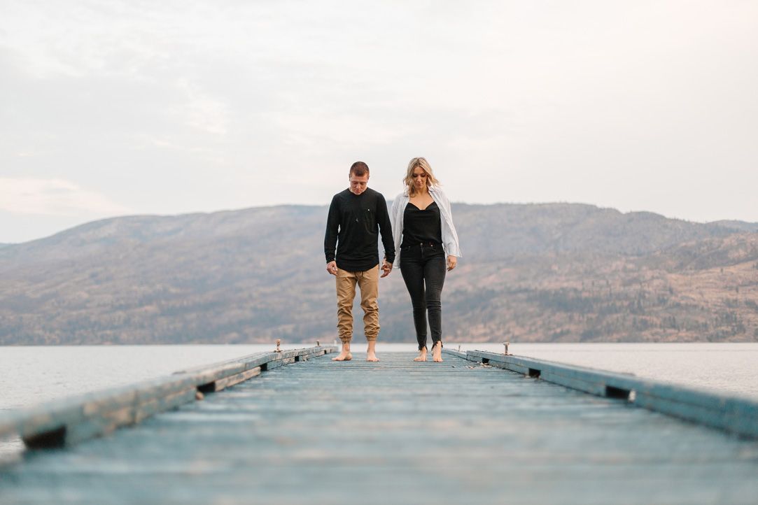 Two people stand at the end of a wooden pier overlooking a mountain lake.