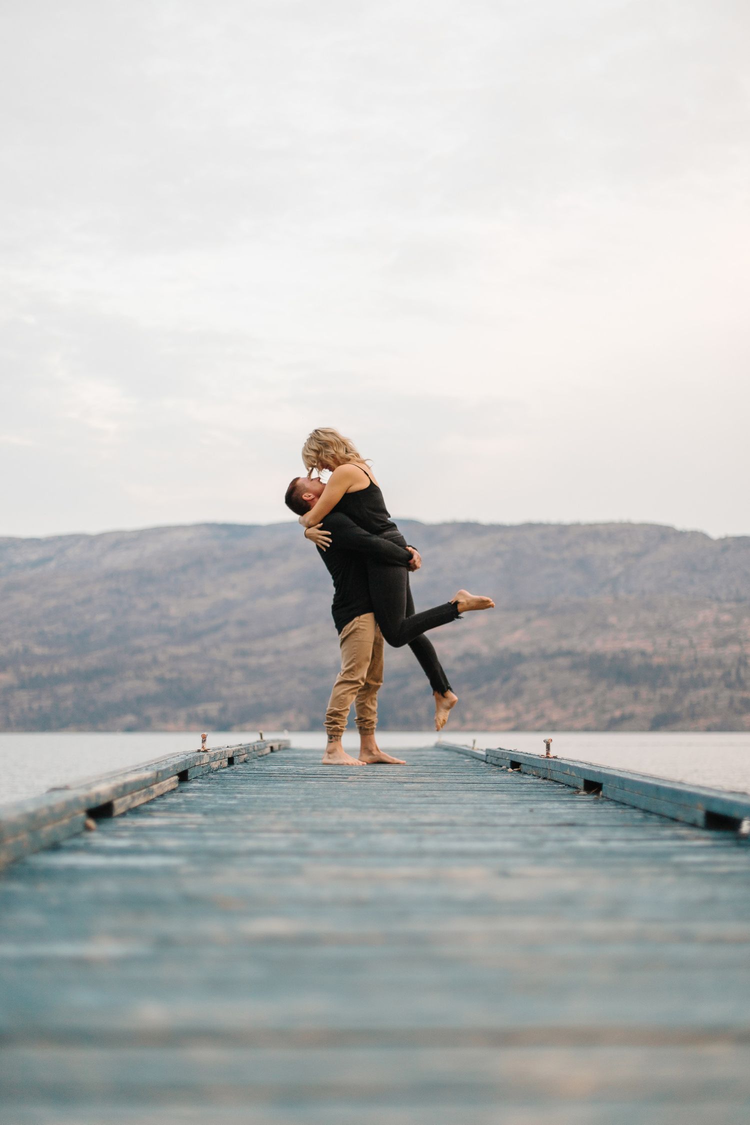Playful couple poses dramatically on a wooden dock overlooking mountain lake.