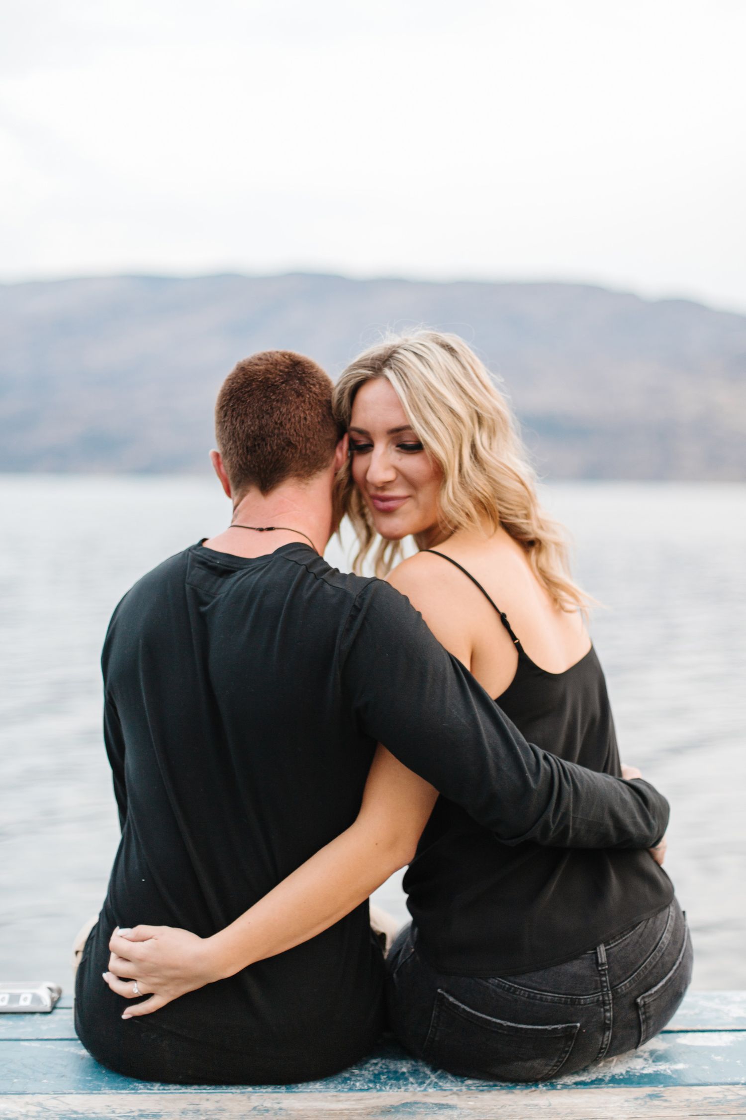 Couple in black outfits sits close together on dock by scenic mountain lake.