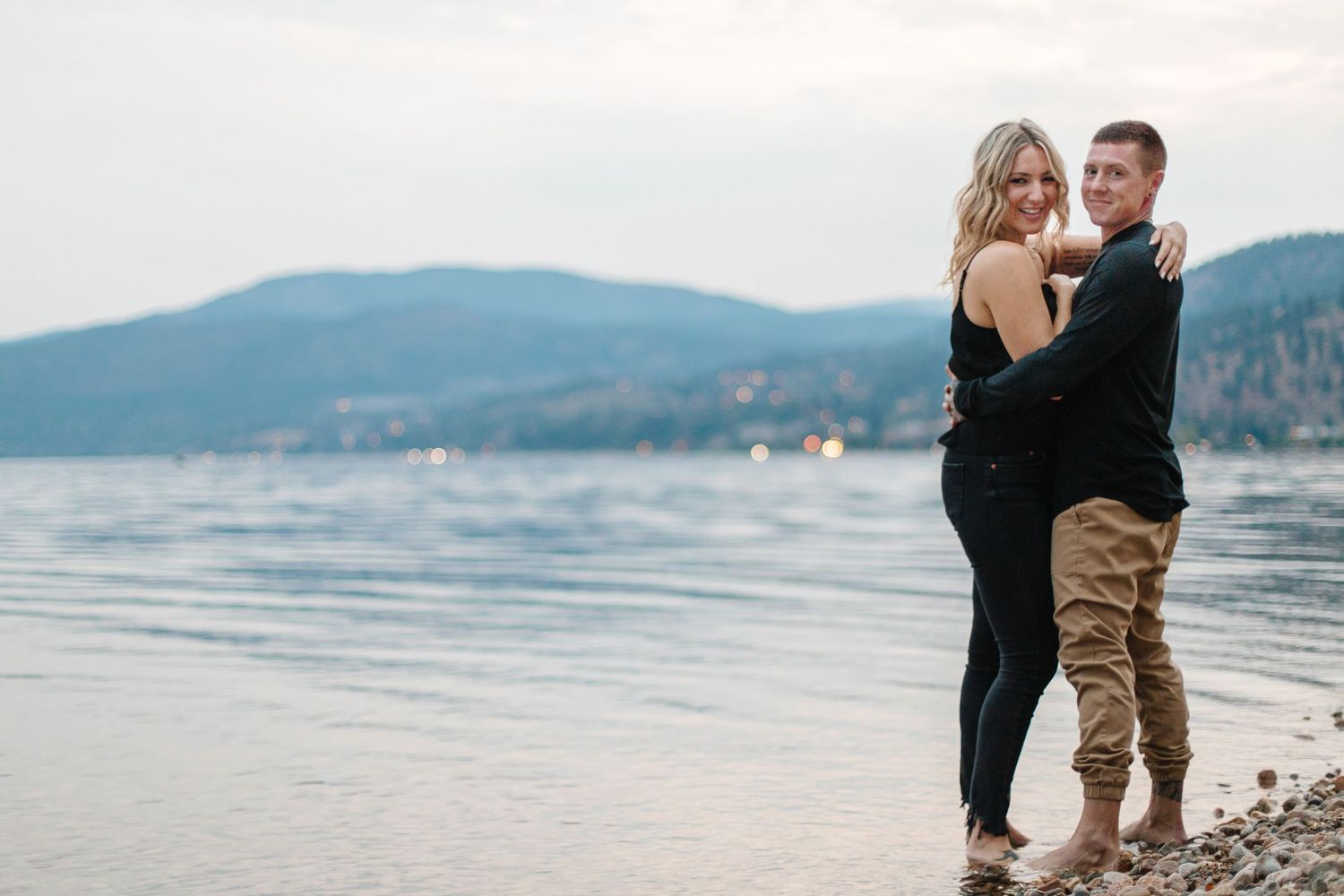 Couple shares intimate moments along rocky lakeshore with mountain backdrop.