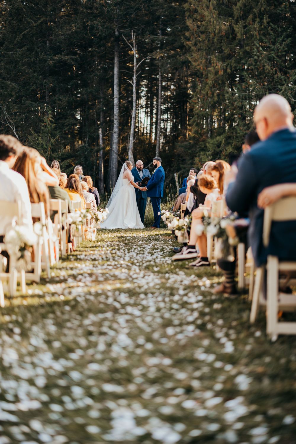 Wedding ceremony aisle lined with white chairs in forest setting with flower petals scattered.