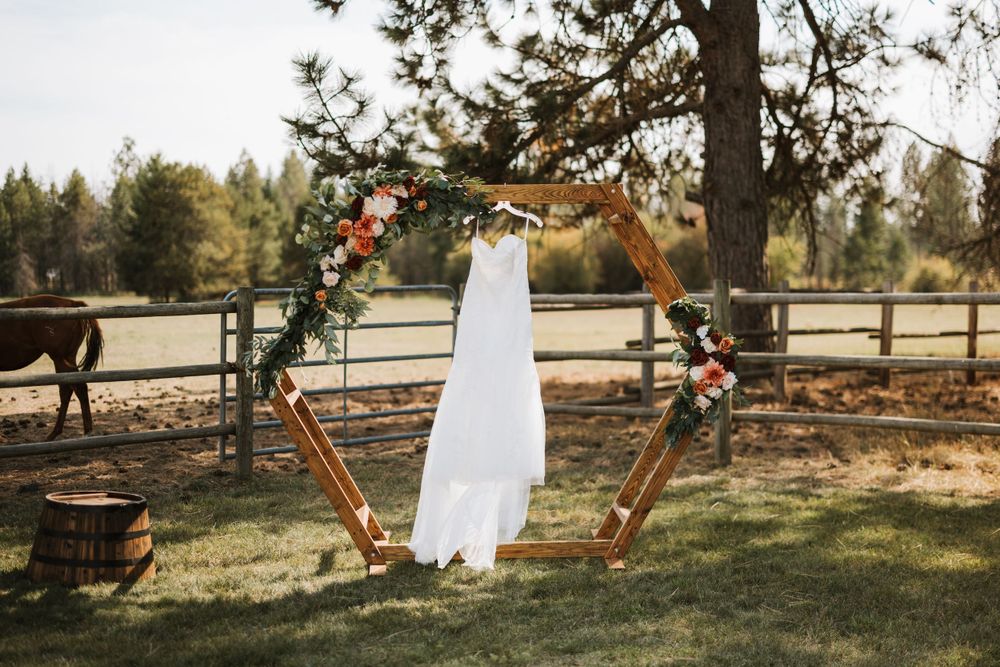 A white wedding dress hangs on a hexagonal copper arch in a rustic outdoor setting with horses nearby.