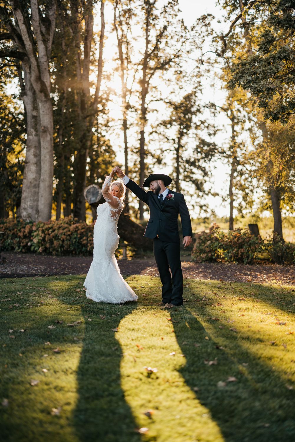 Newlyweds dancing and laughing together in evening sunlight with long shadows on grass.