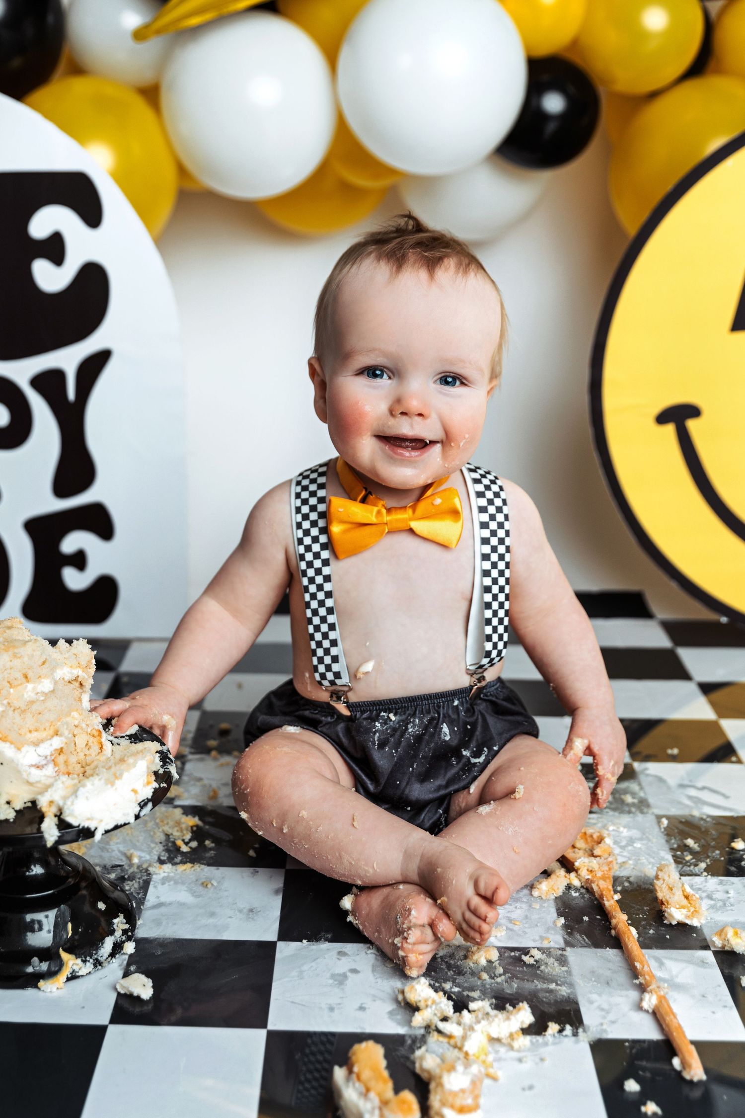 Smiling baby in suspenders and bow tie during cake smash session with yellow balloon decor.