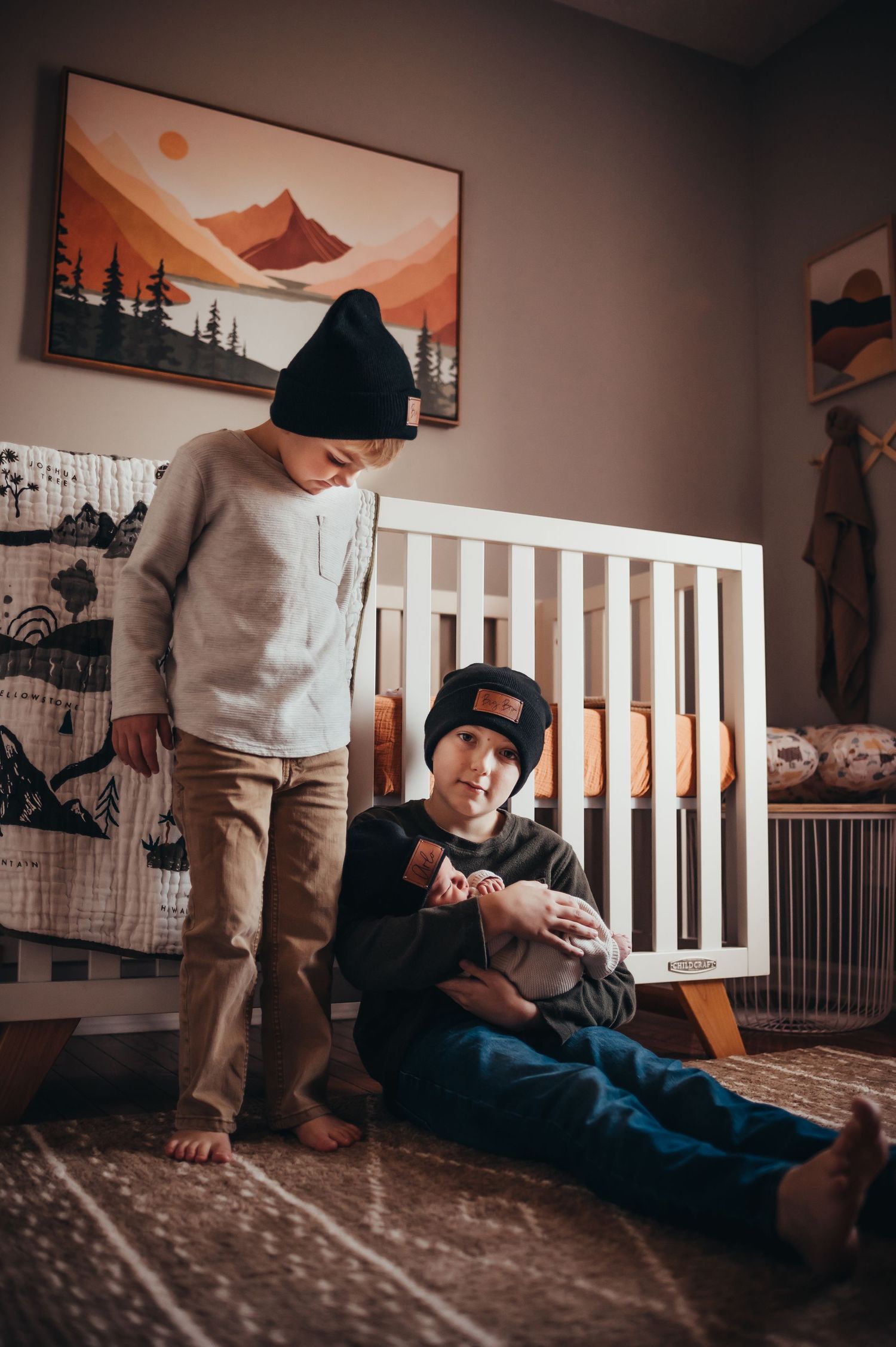 big brother holds newborn little brother in front of the crib while other big brother stands and watches beside them