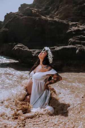 Artistic beach portrait of a person in white dress laying in shallow surf near dark cliffs.