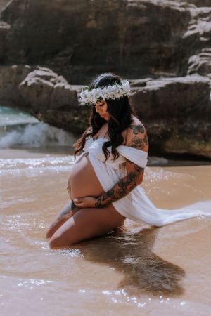 A woman in white poses artistically on a sandy beach near dark rocks wearing a floral crown.