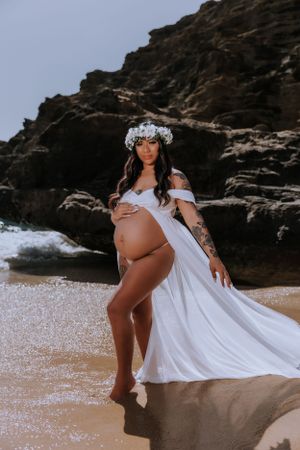 Dramatic beach maternity pose with flowing white dress against coastal rocks.
