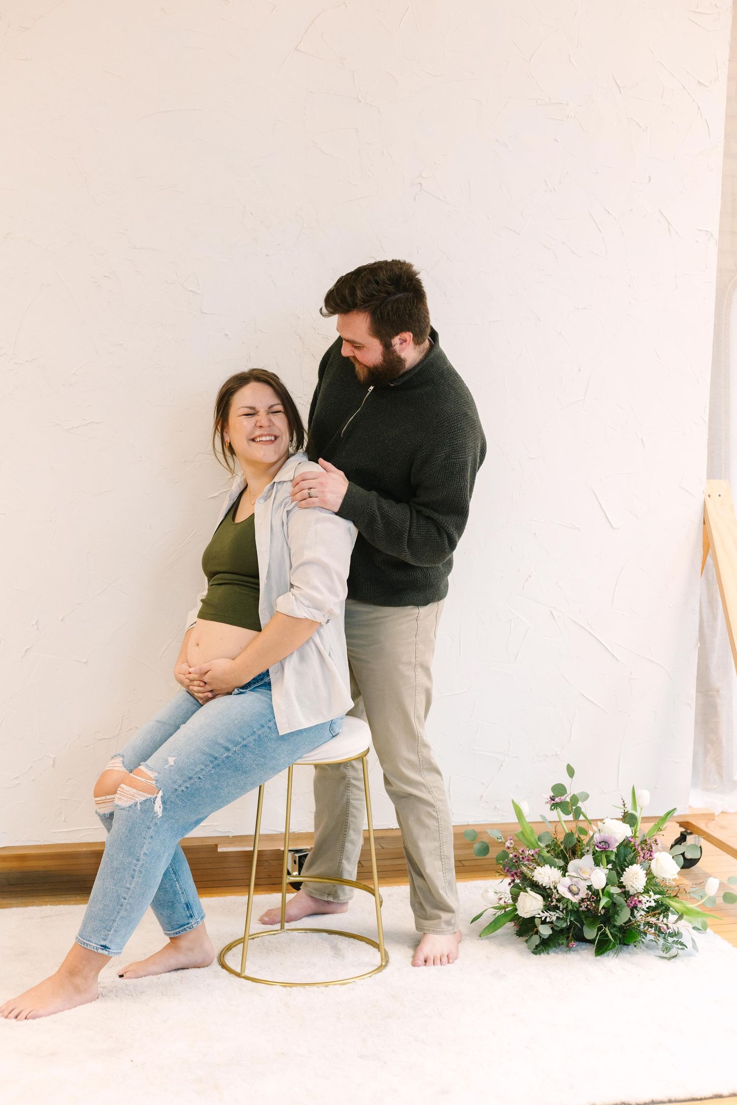 A romantic maternity photo features a couple posing casually with a small floral arrangement on the floor.
