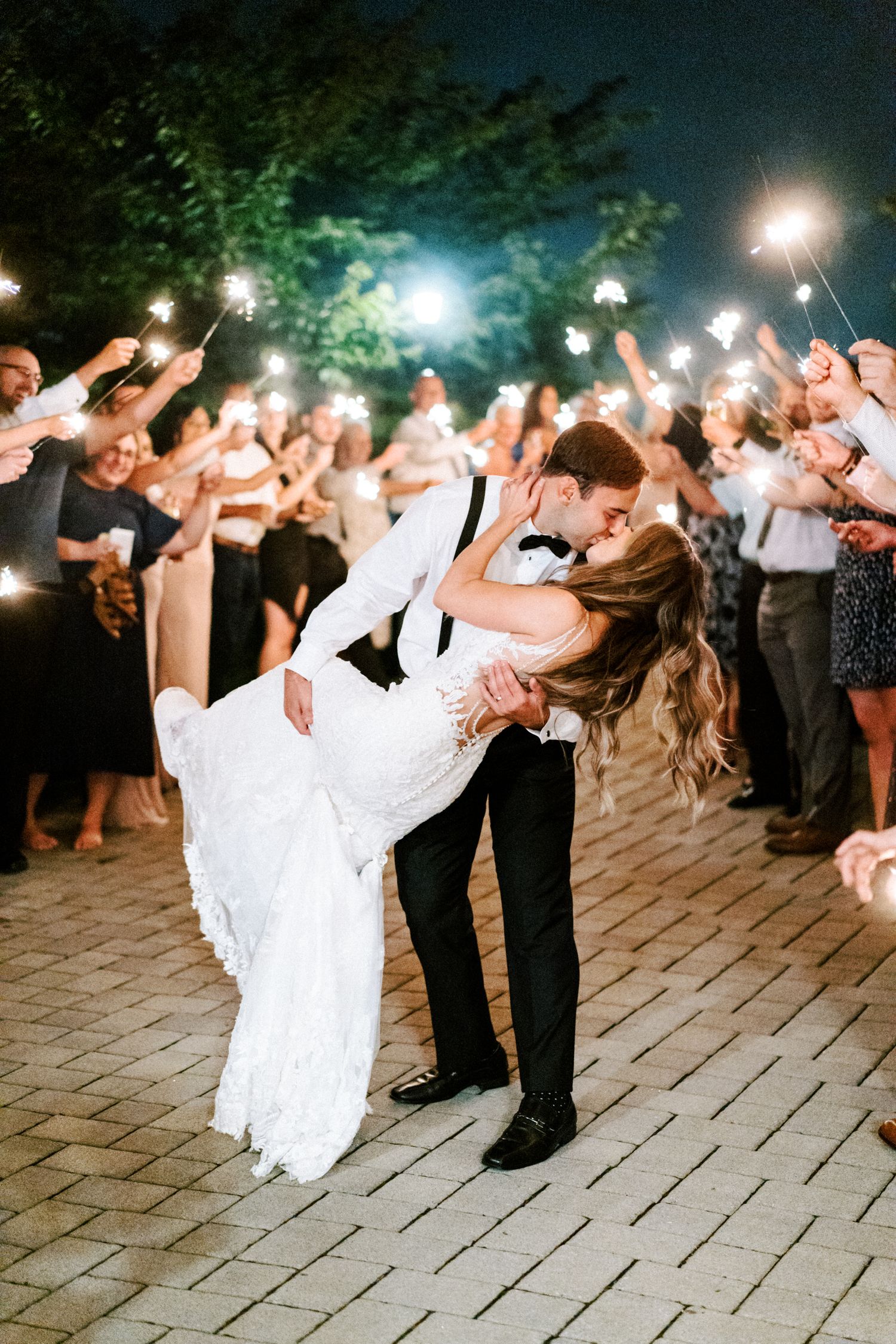 Romantic dance dip under sparklers at an evening wedding celebration on brick pavement.