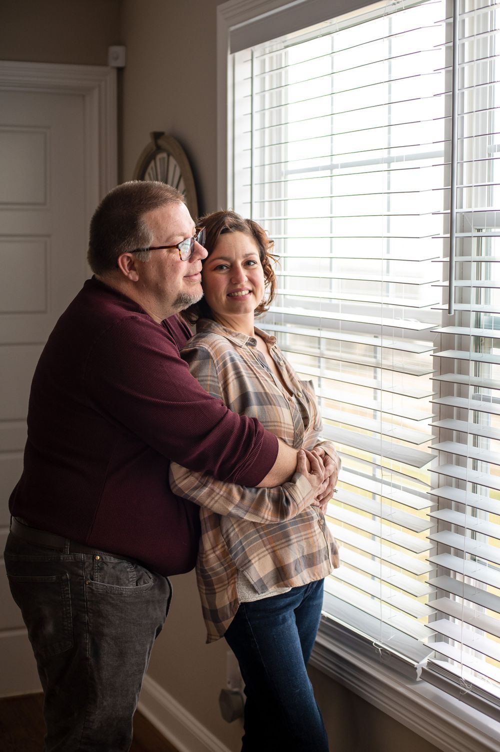 Couple embraces by bright window with venetian blinds in cozy home setting.
