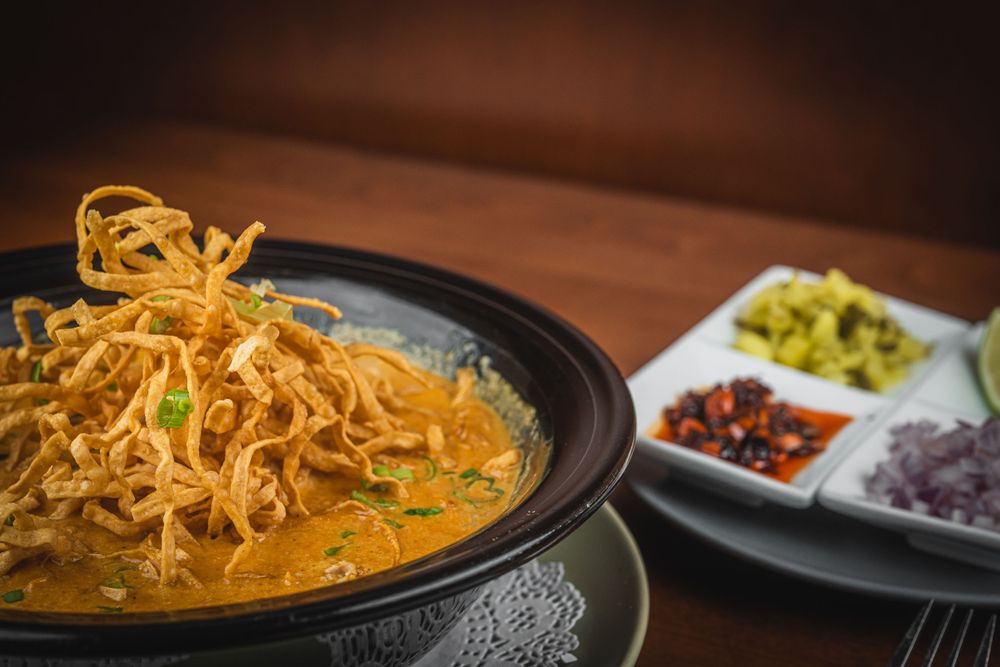 A bowl of Thai khao soi curry noodle soup with crispy noodles and side condiments on a wooden table.