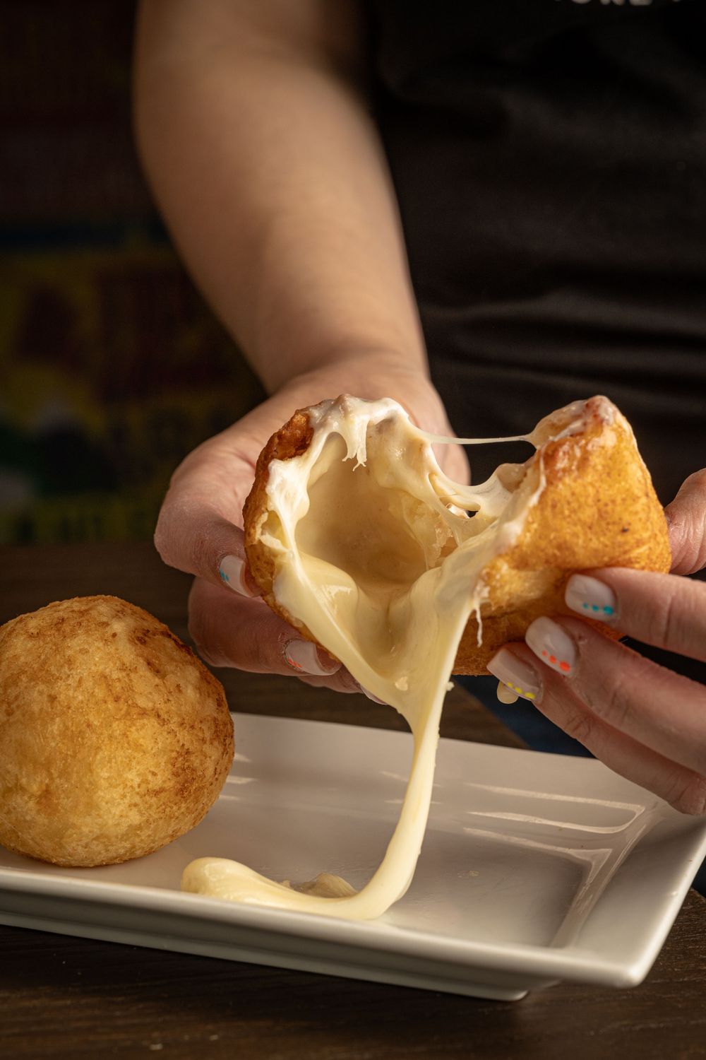 Hands pulling apart a golden fried cheese ball revealing a stretchy melted cheese filling on a white plate.