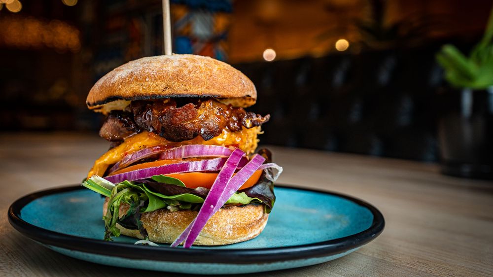 A towering gourmet burger with fresh greens, red onion, and toppings served on a blue plate.