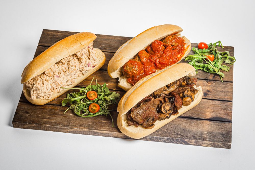 Three gourmet sub sandwiches with toppings on a rustic wooden board.