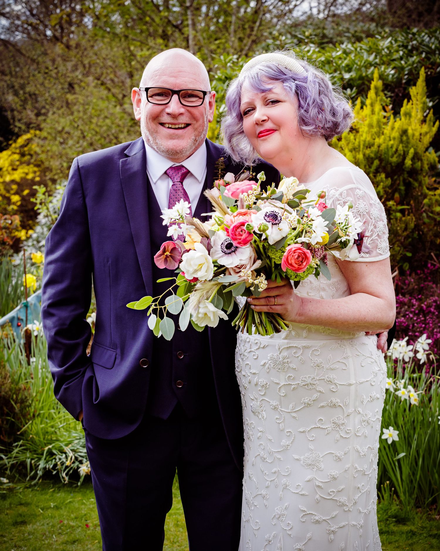 A couple posing in their wedding attire with a vibrant floral bouquet in a garden setting.