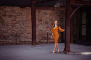 A flowing orange wrap dress with puff sleeves showcased against a rustic brick wall background.