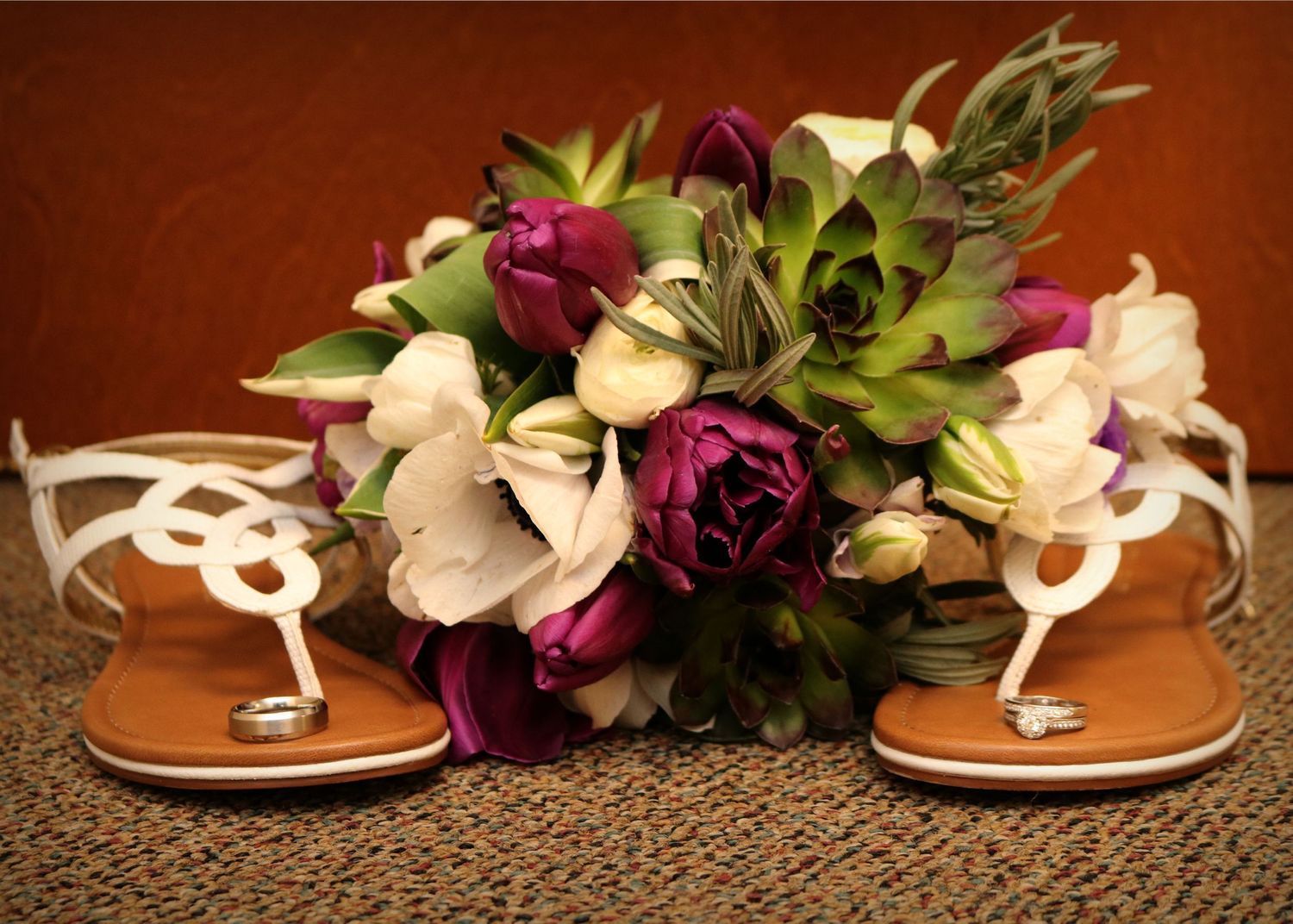White sandals displayed with deep purple and white floral bouquet against brown background.