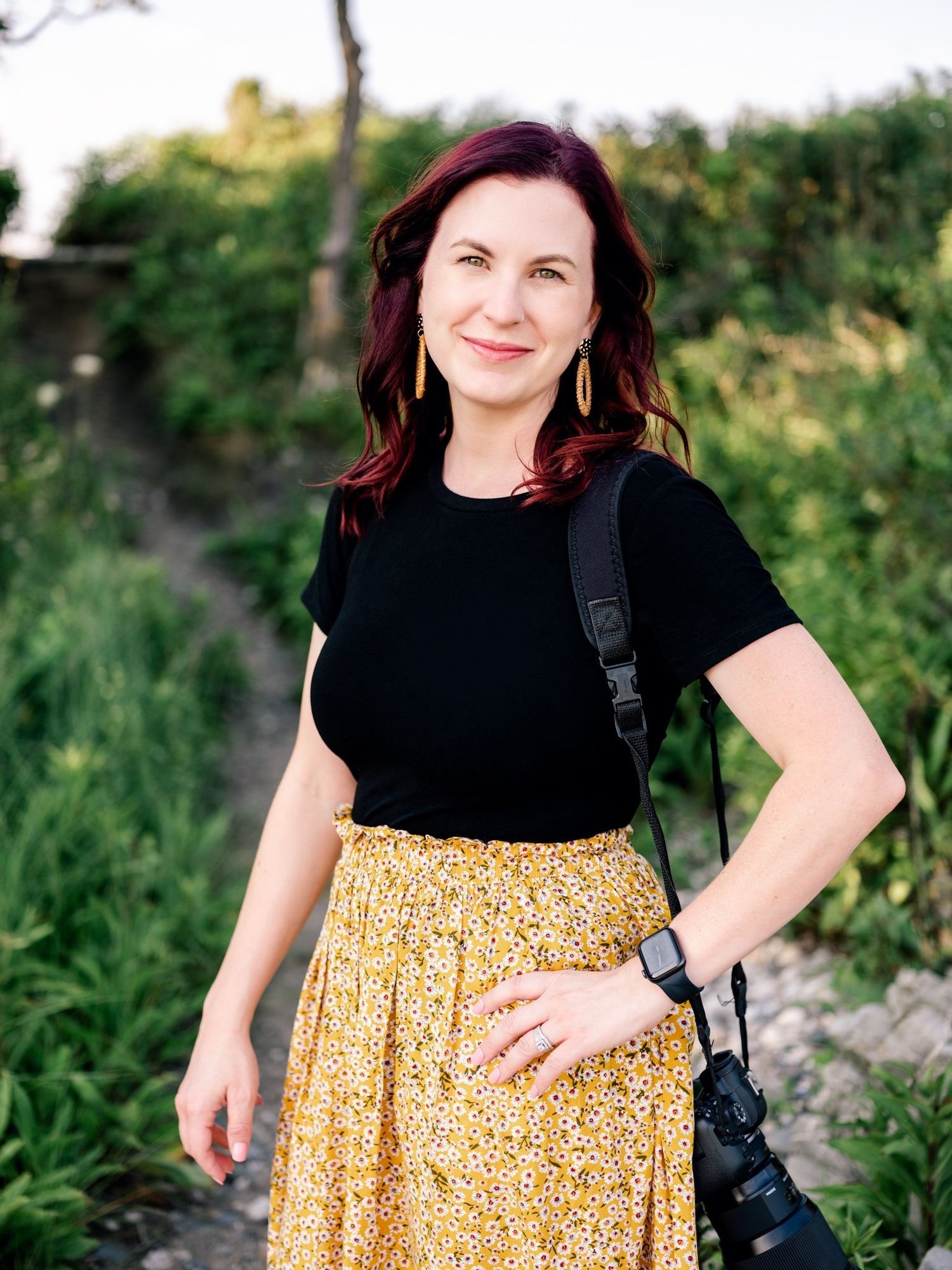 Photographer in black shirt and yellow floral skirt smiling while posing outdoors.