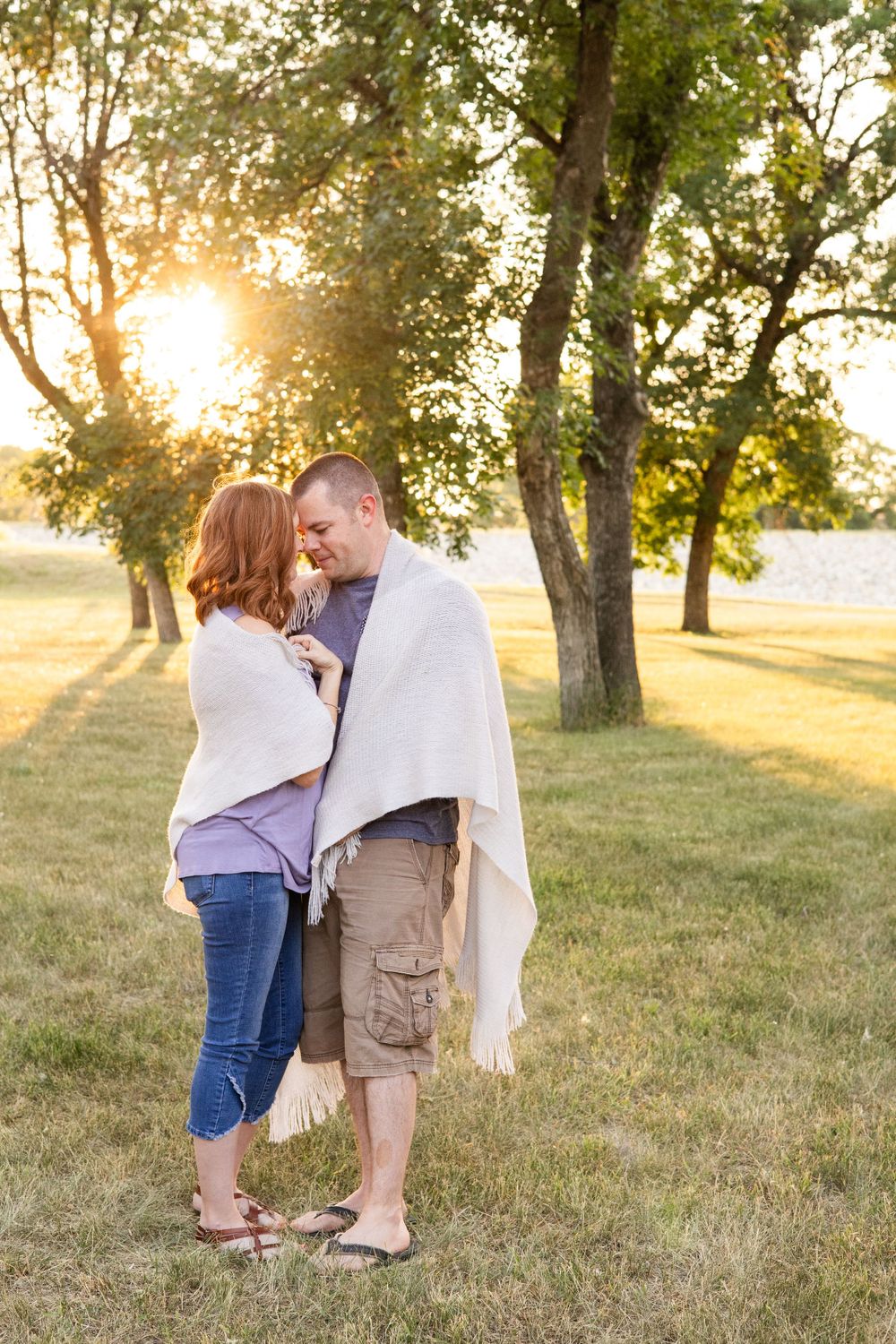 A couple wrapped in a blanket shares an intimate moment at sunset in a park with trees.