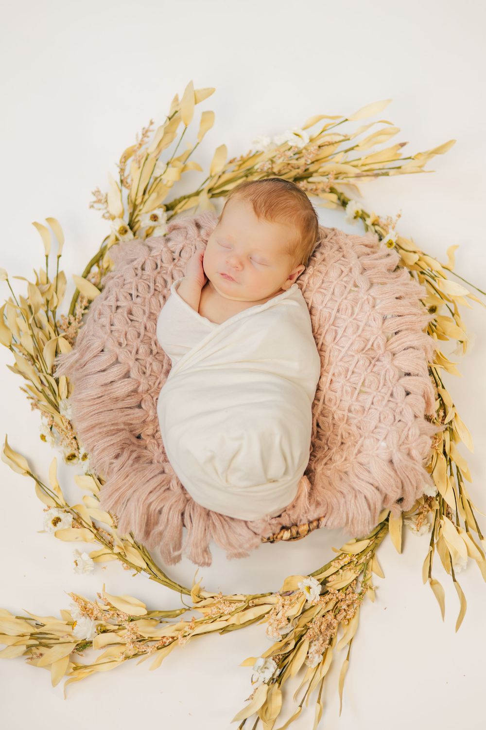 Sleeping newborn wrapped in white swaddle surrounded by dried wheat on pink textured fabric.