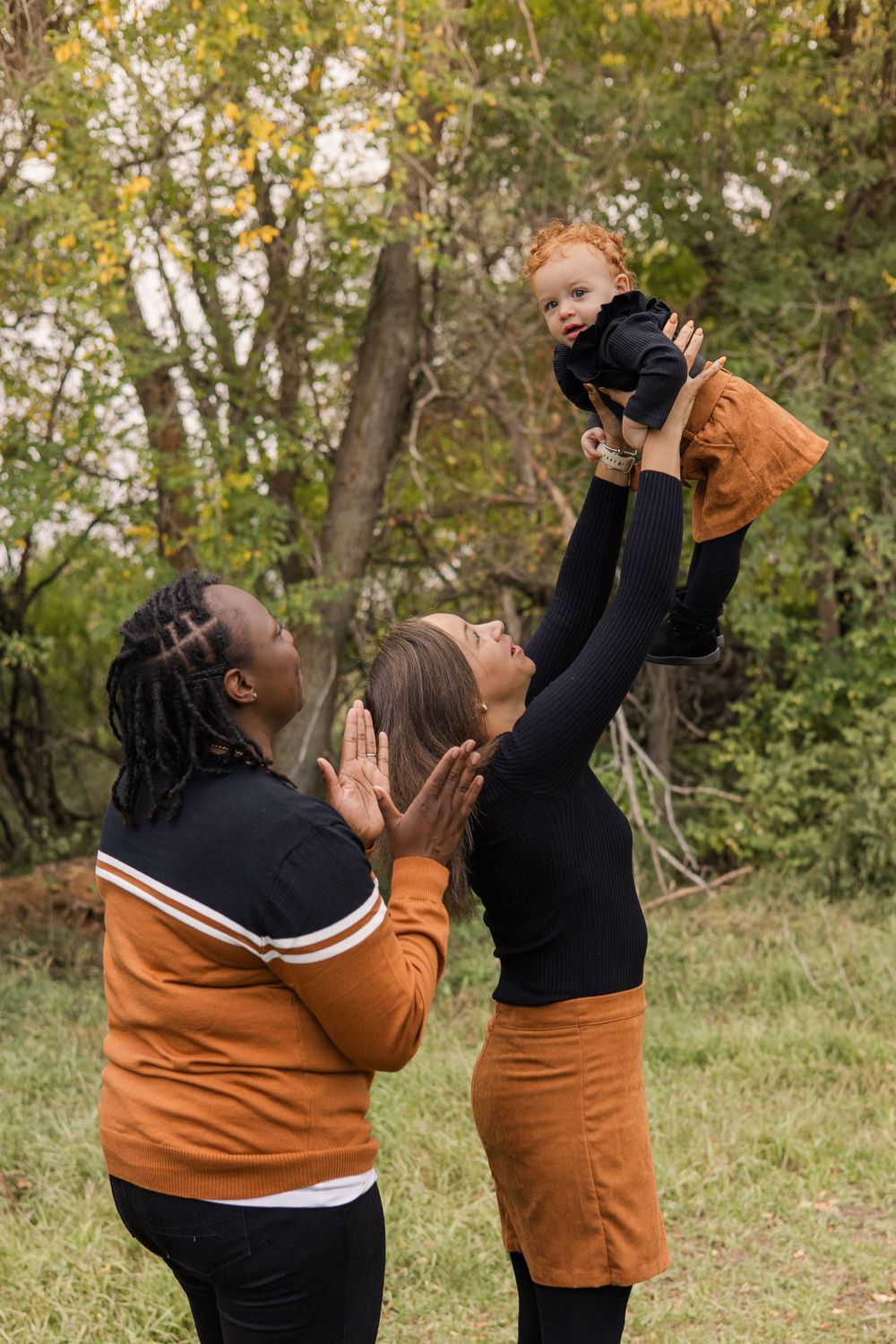 Two people in coordinating orange and black outfits lift and play with a small child outdoors.