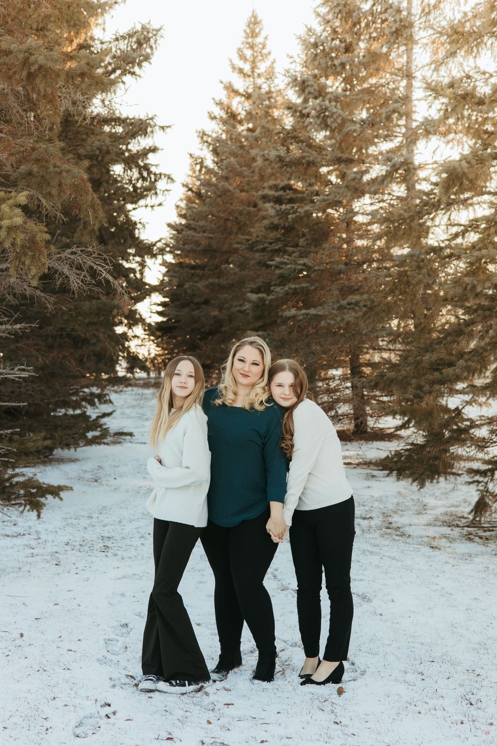 Three people in winter clothing pose together in a snowy setting with evergreen trees.