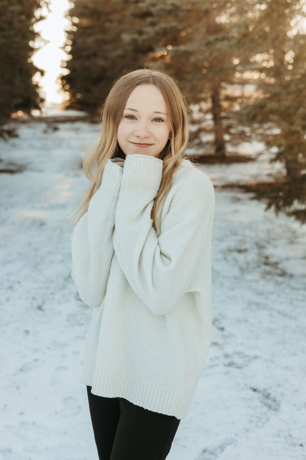 Person in white sweater poses against snowy winter landscape at sunset.